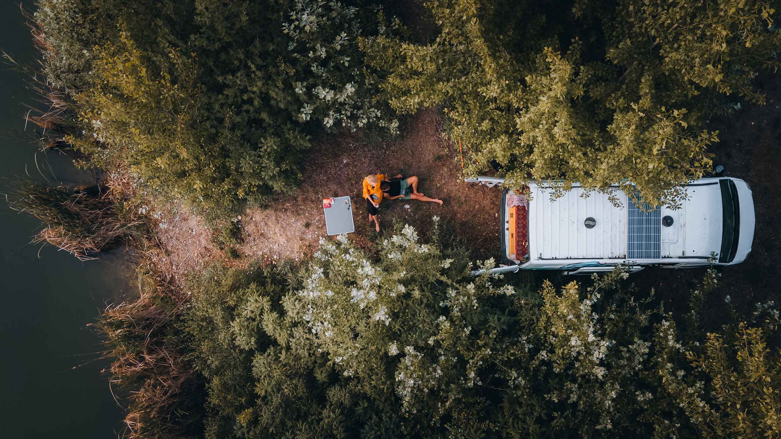 Drohnenaufnahme eines Campervans am Waldrand neben einem See, mit zwei Personen, die am Lagerfeuer sitzen.