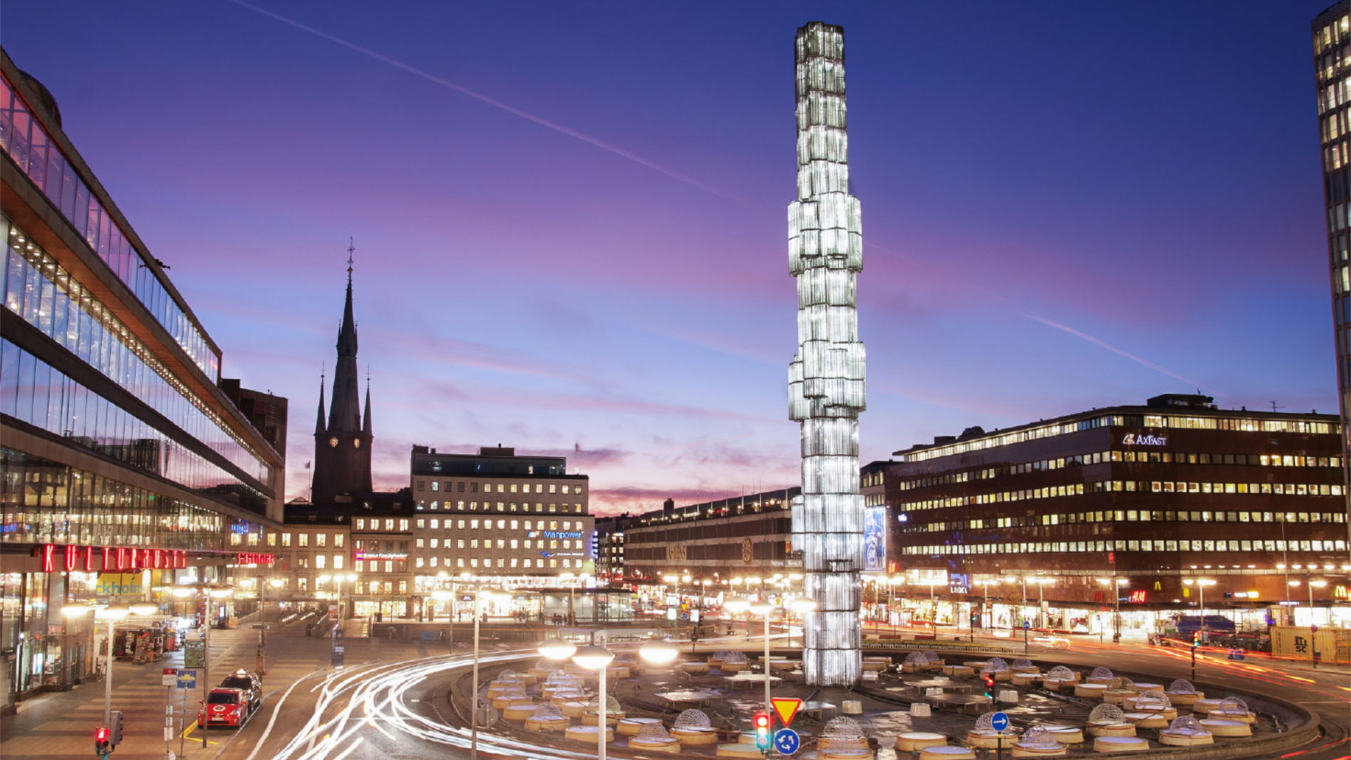 Abendliche Stadtansicht mit beleuchtetem Platz und markantem Glasmonument.