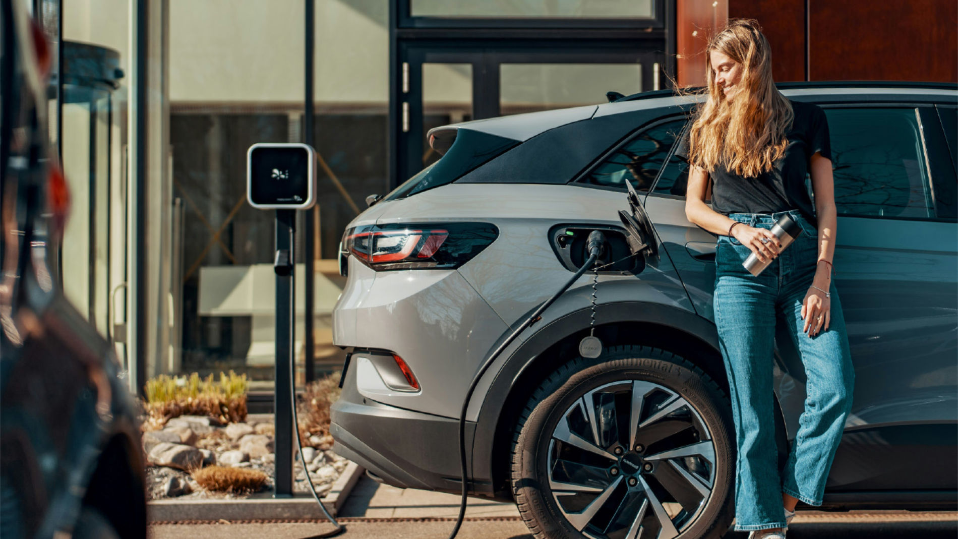Frau lehnt an einem Elektroauto, das an einer Ladestation aufgeladen wird