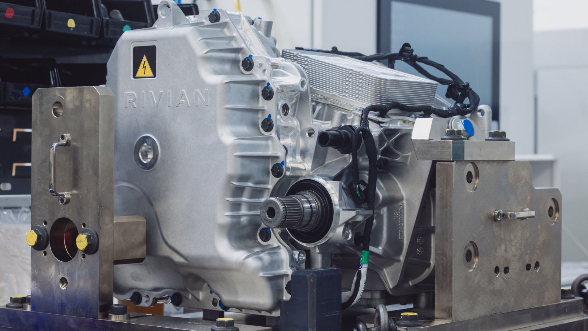 Silver machine part from Rivian with cables and connectors, mounted on a test bench.