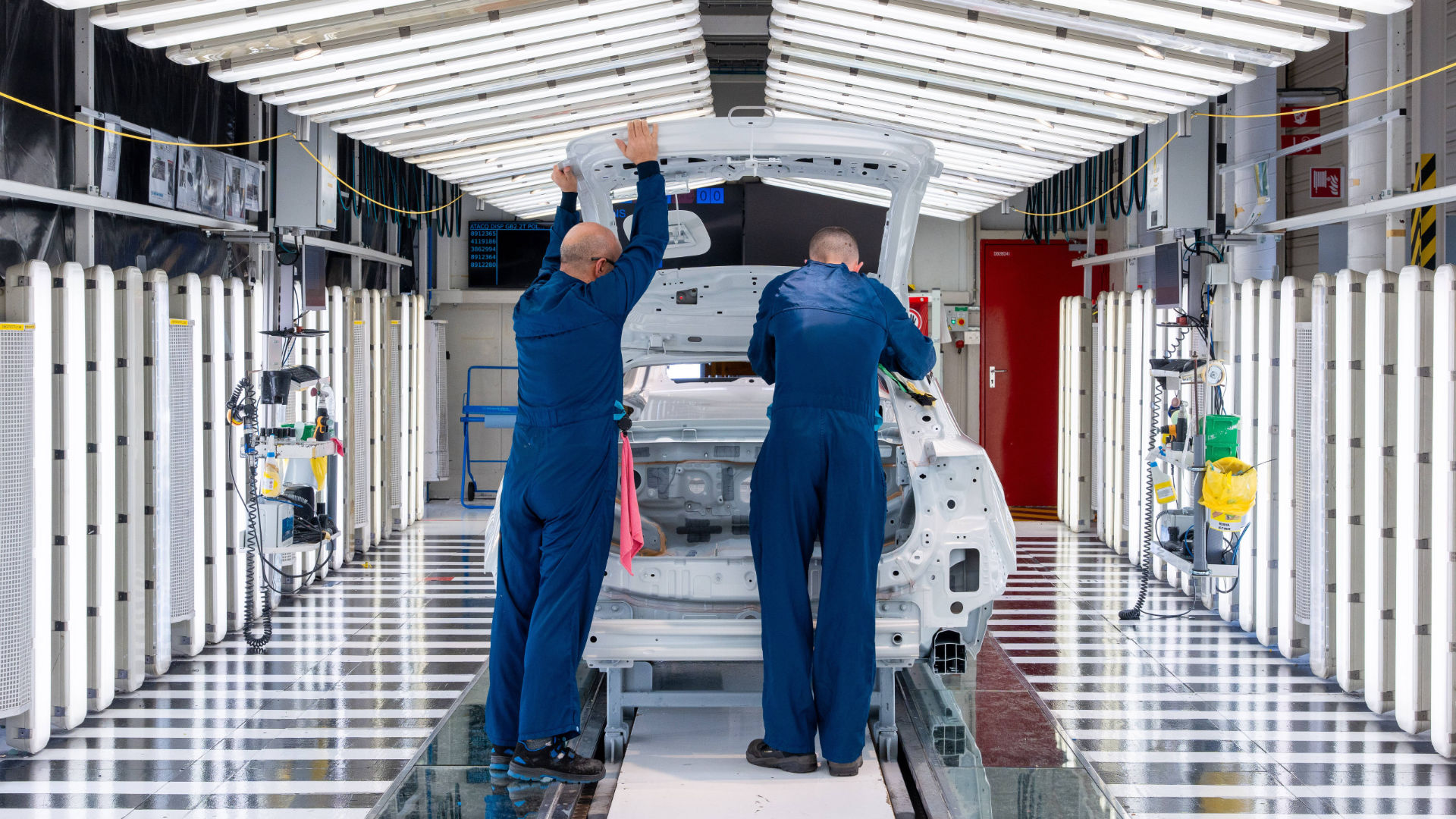 Two workers assembling a car body in a brightly lit production hall.